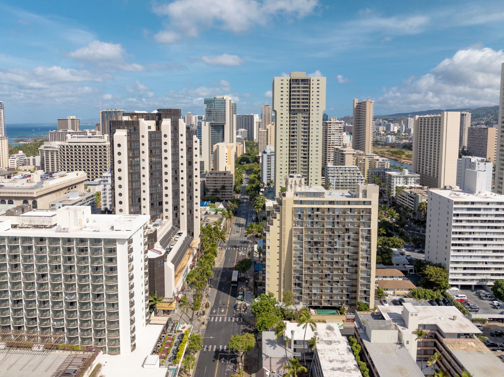 An urban skyline with tall buildings and palm trees line the streets, leading to a distant ocean, under a partly cloudy sky.