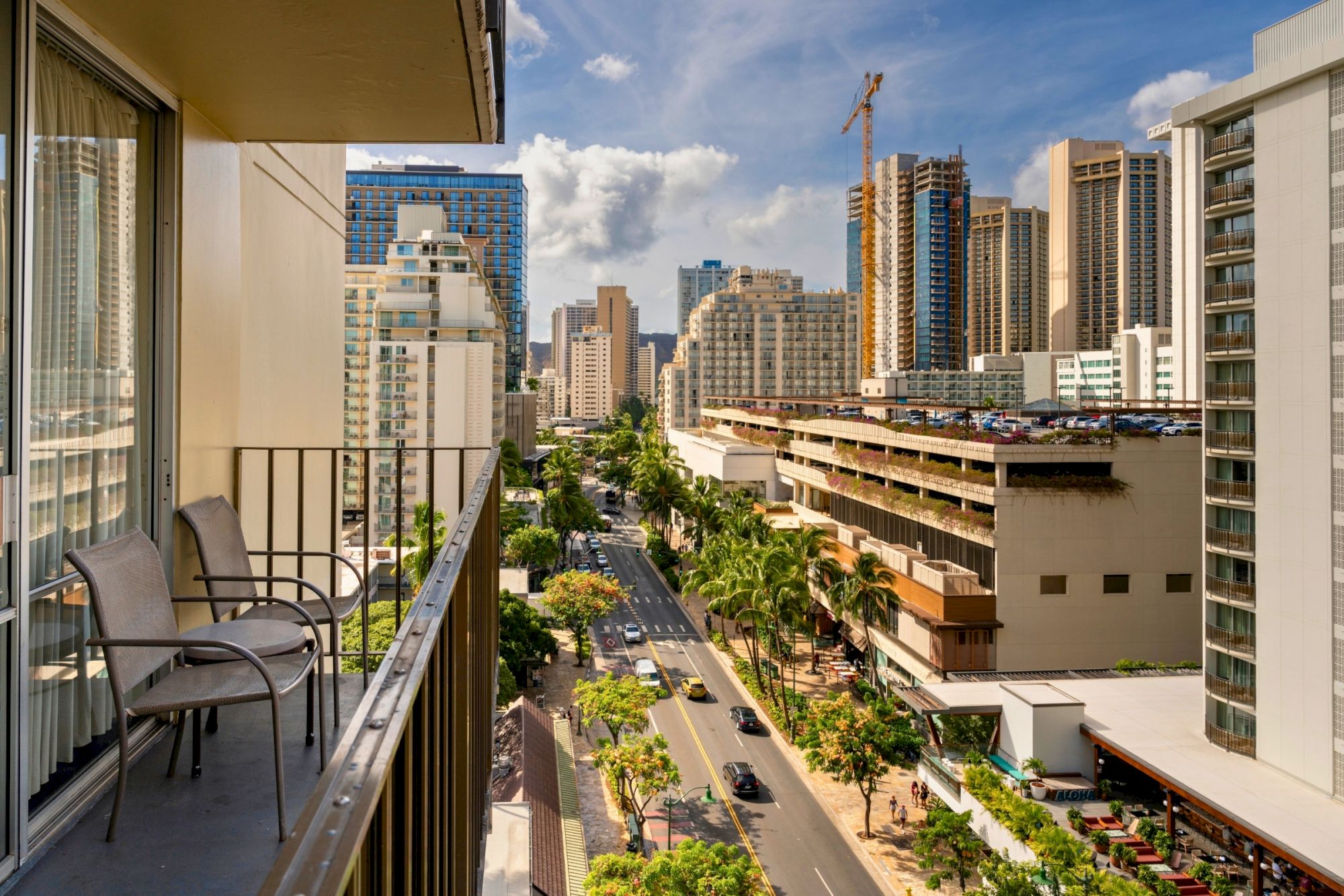 A cityscape view from a balcony, showing a street lined with tall buildings and palm trees under a blue sky with clouds.
