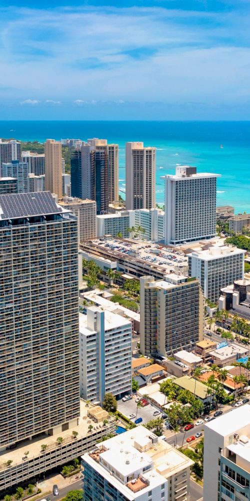 An aerial view of a coastal city with tall buildings, a blue ocean, and a mountain in the background under clear skies.