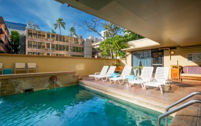 The image shows an outdoor swimming pool with sun loungers, surrounded by buildings and trees under a blue sky.