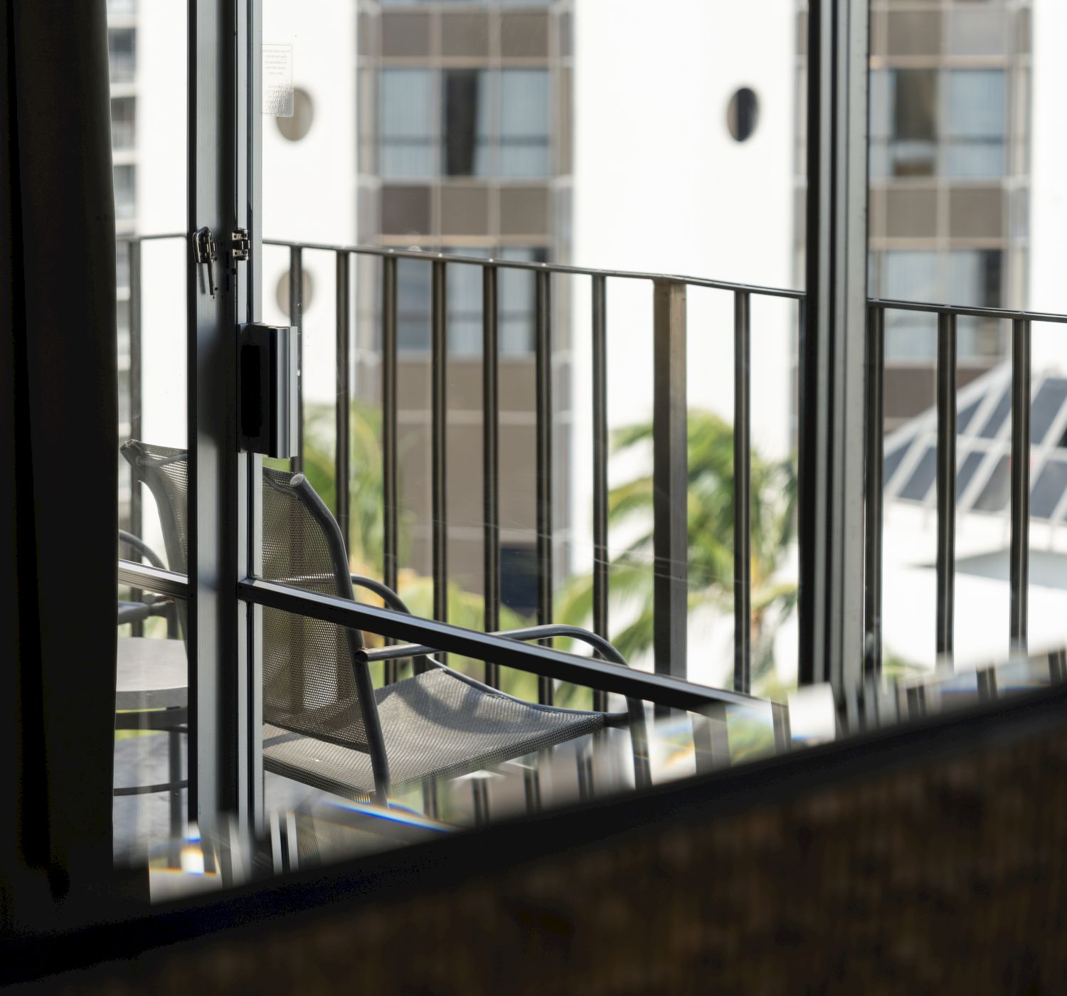 A reflection in a window shows a balcony with chairs, railing, and a view of a building across the way.