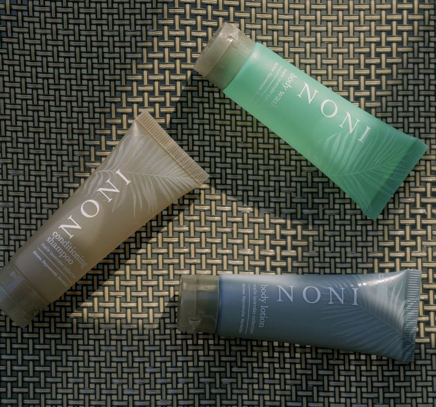 Three tubes of NONI skincare products on a woven surface, with varied colors and sunlit patterns on the tubes.