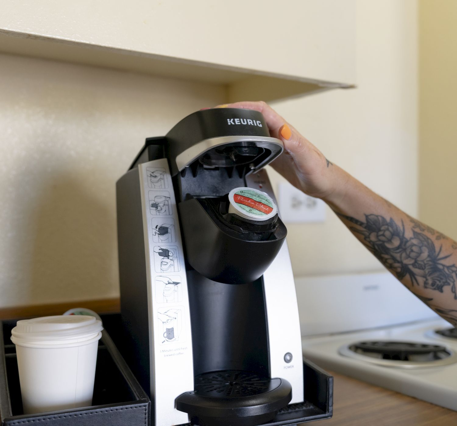 A person using a coffee machine next to a tray with a disposable cup, beside a stove in a kitchen setting.