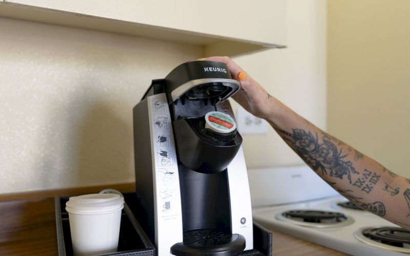 A person's arm with tattoos reaching for a Keurig coffee maker on a counter with a white cup.