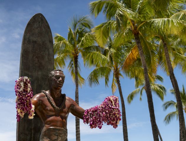 A statue of a surfer with leis is in front of palm trees and a surfboard under a clear blue sky.