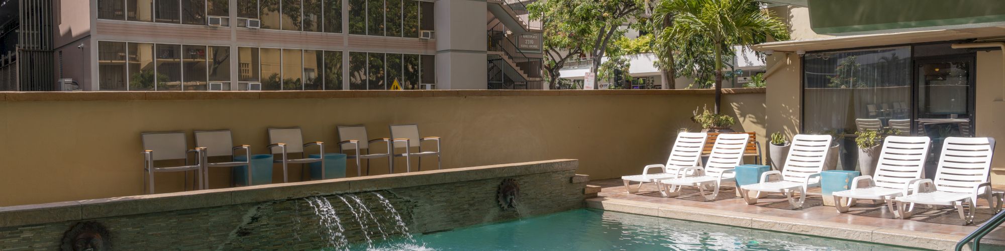The image shows an outdoor pool area with lounge chairs, a waterfall feature, and surrounding buildings casting shadows.