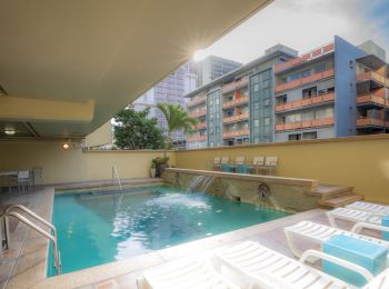 A serene swimming pool area with lounge chairs, modern buildings in the background, and sunlight streaming in from above.