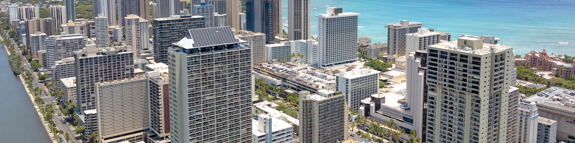 Aerial view of a coastal cityscape with skyscrapers, a canal, and a beach. Ocean and mountains are visible in the background.