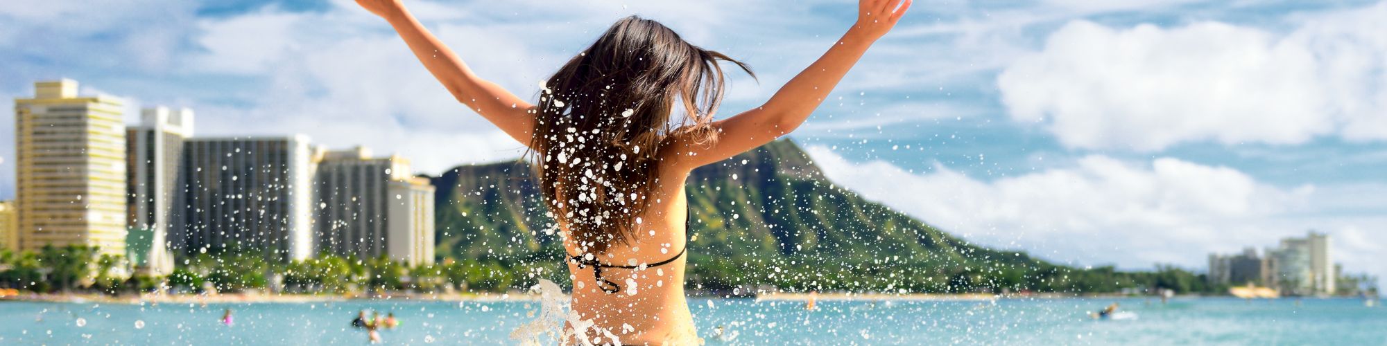 A person in a bikini is joyfully splashing in the water on a sandy beach with city buildings and a mountain in the background, under a clear sky.