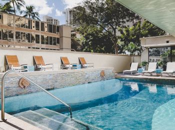 The image shows an outdoor swimming pool with lounge chairs, surrounded by buildings and trees, under a partly cloudy sky.