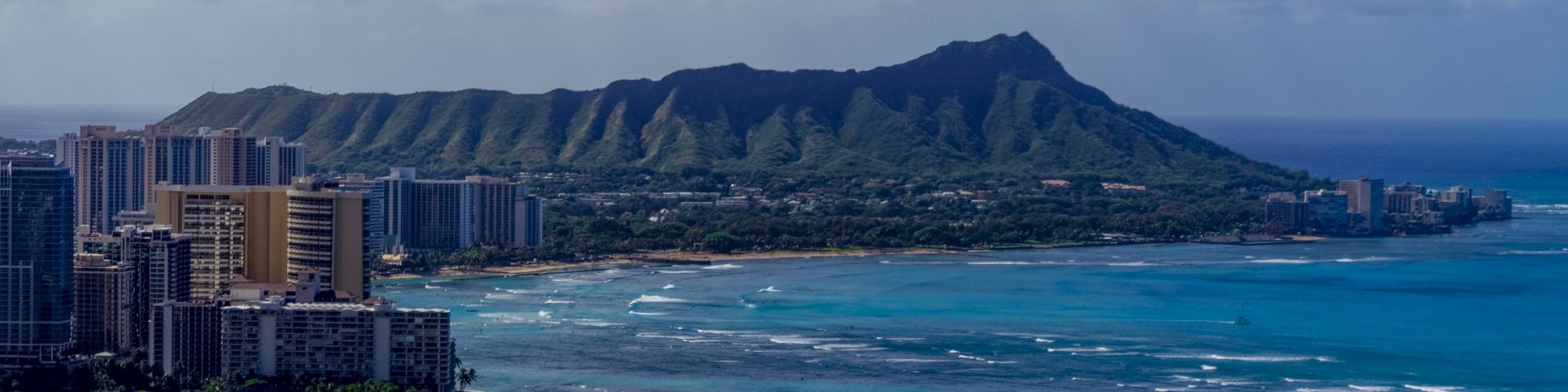 A coastal cityscape with sandy beaches, turquoise ocean, and a mountainous backdrop under a partly cloudy sky.