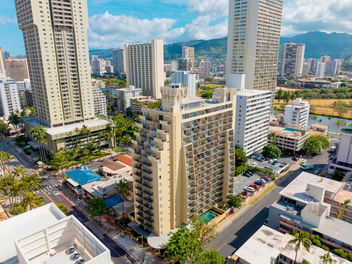 An aerial view of a cityscape featuring several tall buildings with a backdrop of mountains and partly cloudy skies.
