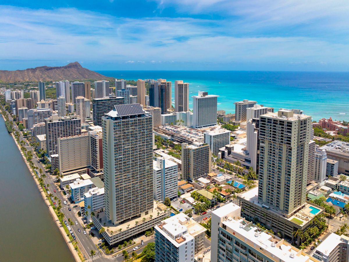Aerial view of a coastal cityscape with high-rise buildings, blue ocean, and a distant mountain under a clear blue sky.