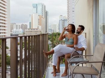 A couple is relaxing on a balcony with a city view; one is using a phone, and the other is sipping from a cup, surrounded by tall buildings.