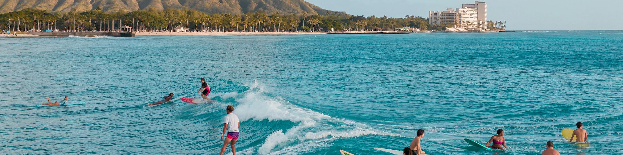 Surfers enjoy the waves with a scenic backdrop of mountains and a city skyline under a clear blue sky.