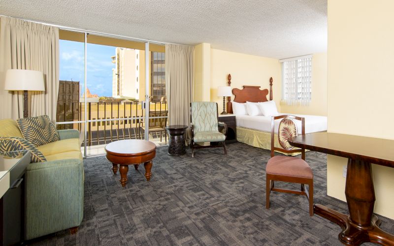 A wide view of a hotel suite with a sofa, coffee table, armchair, desk, bed, and balcony with city view.