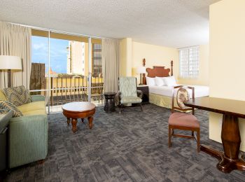 The image shows a hotel room with a seating area, bed, and balcony. There are chairs, a sofa, a table, and curtains.