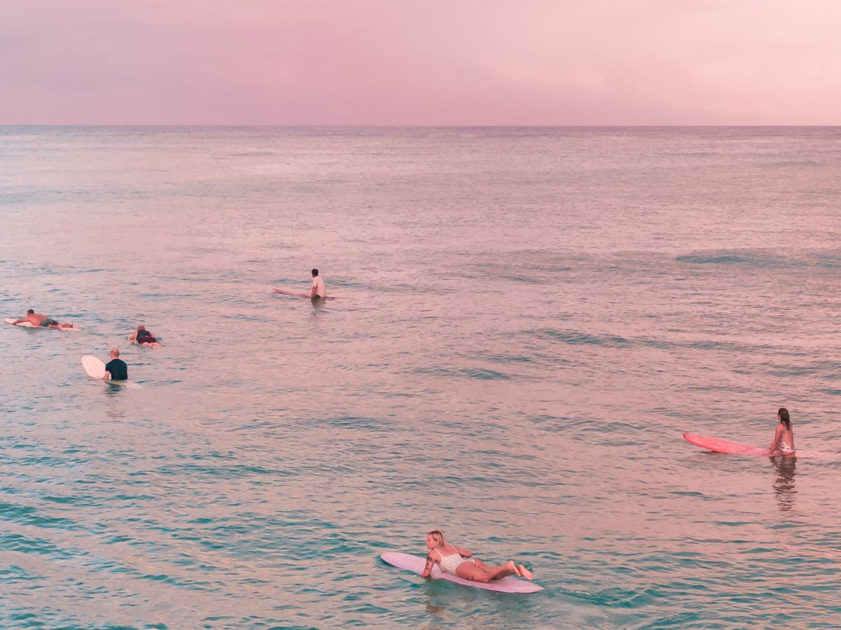 People on surfboards are floating on calm ocean waters under a pinkish sky during sunset.