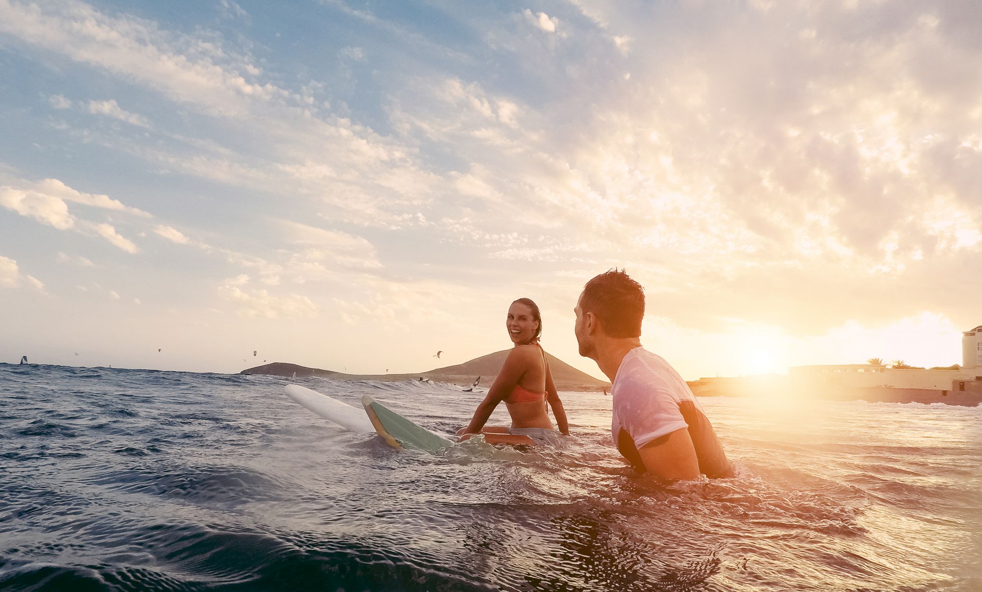 Two people in the ocean preparing to surf, with one holding a surfboard, under a beautiful sunset sky.