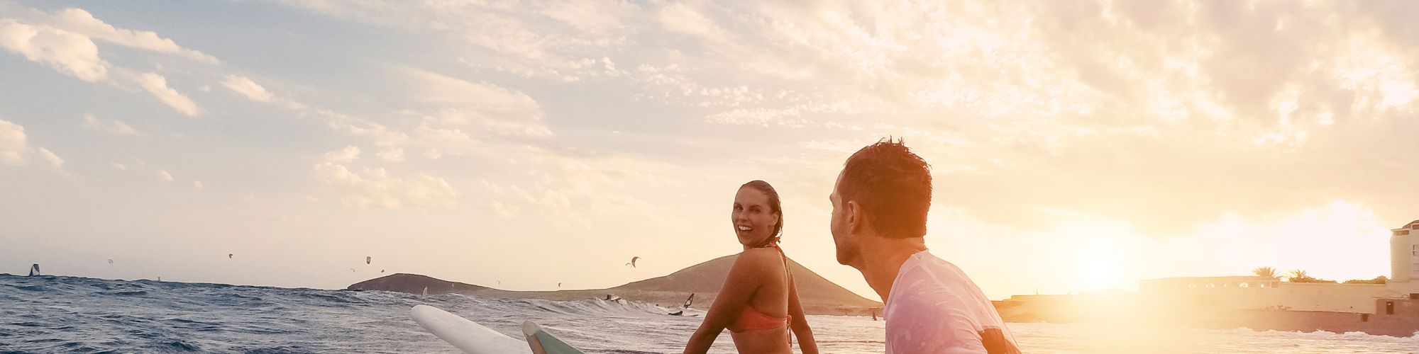 Two people in the ocean preparing to surf, with one holding a surfboard, under a beautiful sunset sky.