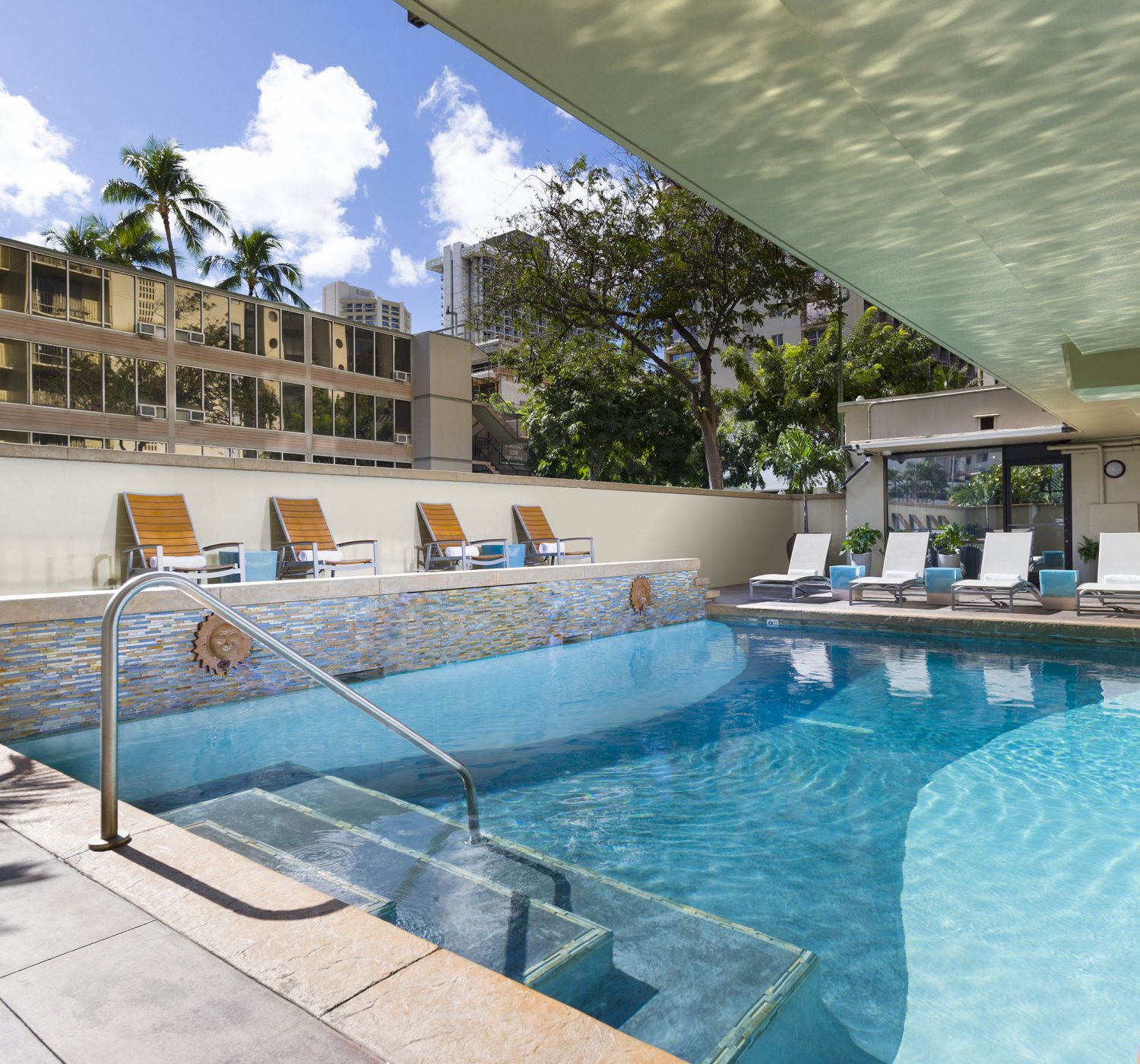 The image shows an outdoor pool with clear water, surrounded by lounge chairs, and a building in the background under a partly cloudy sky.