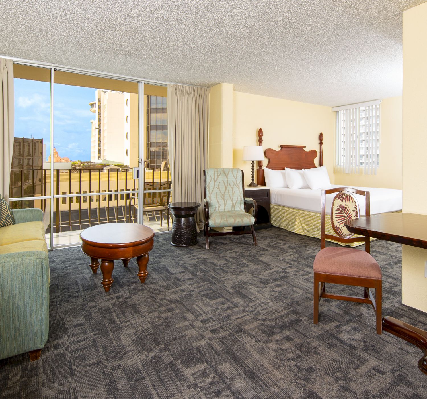 The image shows a hotel room with a living area, sofa, chairs, coffee table, bed, and balcony, all in a light, neutral color scheme.
