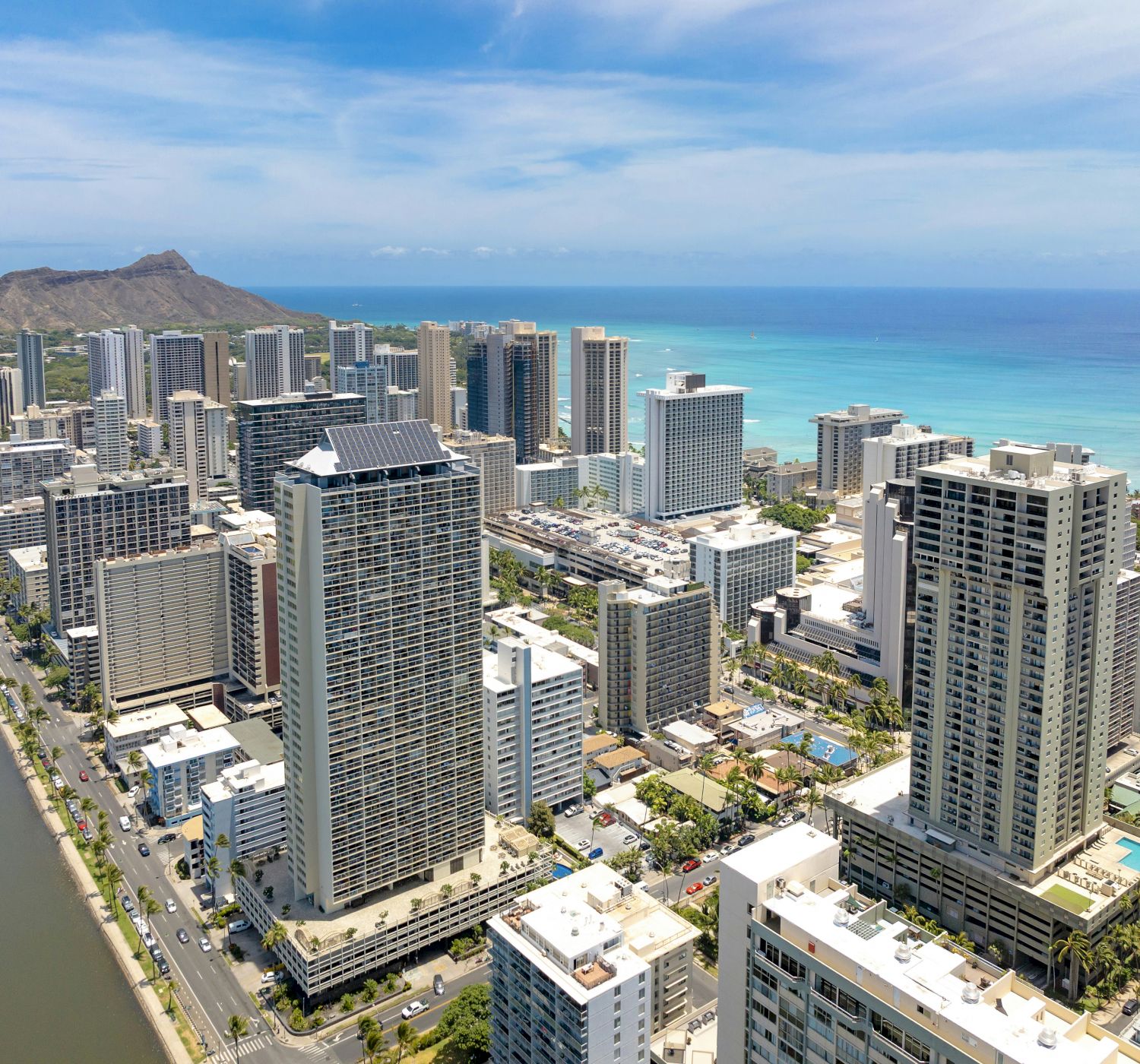 The image shows a cityscape with tall buildings near the ocean, a canal, and a mountain in the background under a clear blue sky.