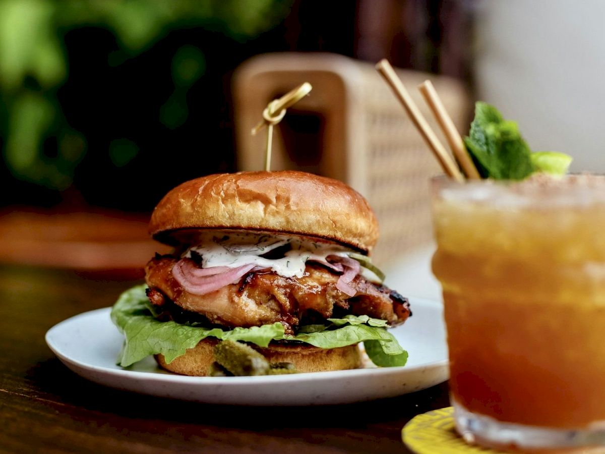 A chicken sandwich with lettuce and onions on a plate, next to a cocktail with lime garnish on a wooden table.