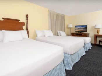 The image shows two neatly made beds in a hotel room with a TV, lamps, and a side table for a comfortable stay.