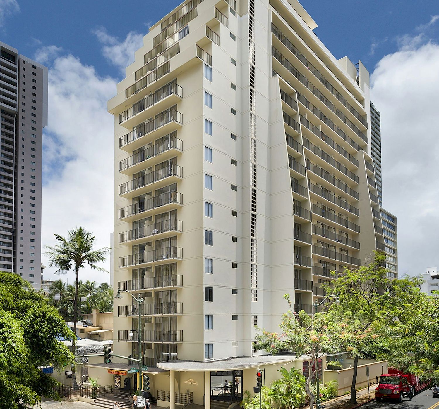 The image shows a tall, modern apartment building with balconies set against a partly cloudy sky, surrounded by trees and a few other buildings.