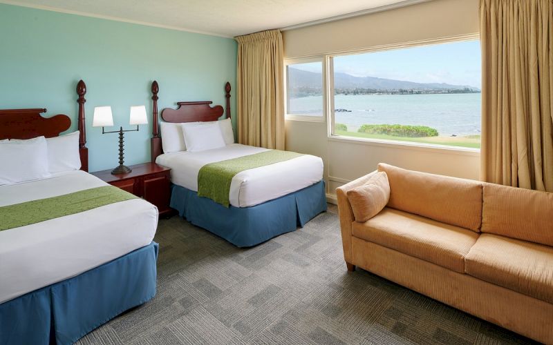 A hotel room with two beds, a sofa, and a large window overlooking the ocean.