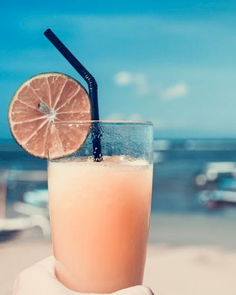 A hand holding a tropical drink with a black straw and a lime slice garnish, against a backdrop of a beach with boats in the water and clear skies.