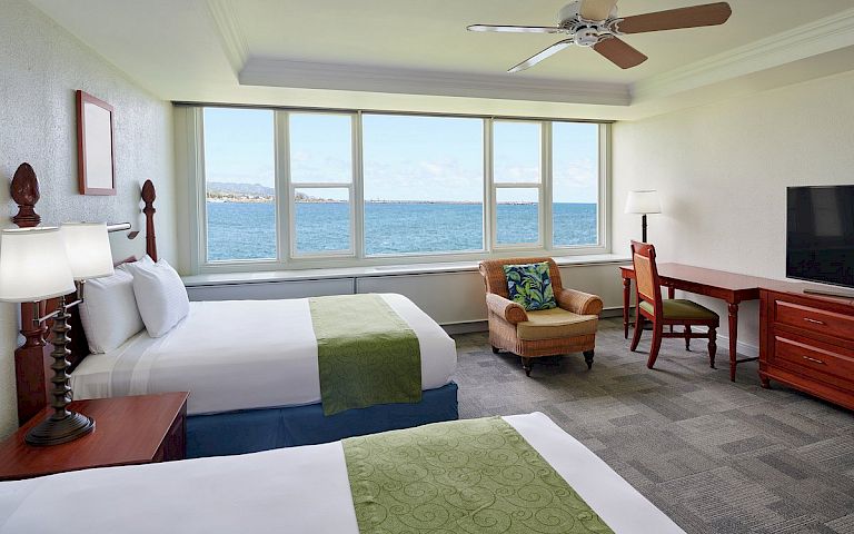 A hotel room with two beds, a desk, and a chair, overlooking the ocean.