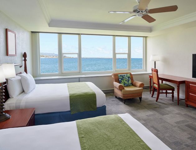 A hotel room with two double beds, a desk, chair, and ocean view.